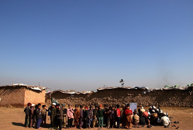 A group of children is gathered in an open area with an expansive blue sky above them. The children are dressed in various layers of clothing, suggesting cooler weather. Behind them, there are low, makeshift mud-brick structures, indicating a rural or underdeveloped setting. Some children are standing, while others are seated on the ground, possibly engaged in an activity.
