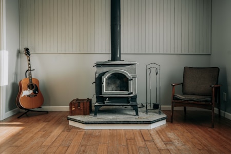 A cozy room with a wooden floor features a black wood-burning stove as the centerpiece. To the left of the stove, an acoustic guitar rests against the wall. On the right side, a vintage wooden chair with fabric cushioning is placed. A small wooden box sits on the floor near the stove, and there are metal fireplace tools beside it. The walls are painted a light color and are adorned with vertical wooden paneling.