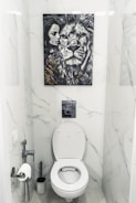 A bathroom with white marble walls and a modern toilet setup. Above the toilet hangs a striking black and white painting featuring a close-up of a lion's face next to a woman's face. The area contains a toilet paper holder with a roll, a toilet brush, and a small handheld bidet.