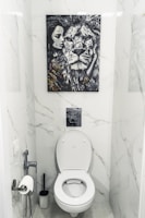 A bathroom with white marble walls and a modern toilet setup. Above the toilet hangs a striking black and white painting featuring a close-up of a lion's face next to a woman's face. The area contains a toilet paper holder with a roll, a toilet brush, and a small handheld bidet.