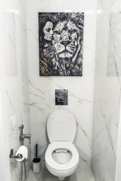 A bathroom with white marble walls and a modern toilet setup. Above the toilet hangs a striking black and white painting featuring a close-up of a lion's face next to a woman's face. The area contains a toilet paper holder with a roll, a toilet brush, and a small handheld bidet.