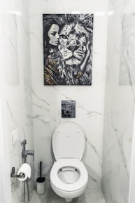 A bathroom with white marble walls and a modern toilet setup. Above the toilet hangs a striking black and white painting featuring a close-up of a lion's face next to a woman's face. The area contains a toilet paper holder with a roll, a toilet brush, and a small handheld bidet.