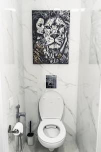 A bathroom with white marble walls and a modern toilet setup. Above the toilet hangs a striking black and white painting featuring a close-up of a lion's face next to a woman's face. The area contains a toilet paper holder with a roll, a toilet brush, and a small handheld bidet.