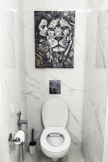A bathroom with white marble walls and a modern toilet setup. Above the toilet hangs a striking black and white painting featuring a close-up of a lion's face next to a woman's face. The area contains a toilet paper holder with a roll, a toilet brush, and a small handheld bidet.