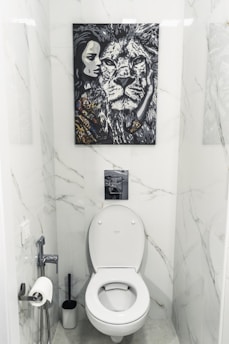 A bathroom with white marble walls and a modern toilet setup. Above the toilet hangs a striking black and white painting featuring a close-up of a lion's face next to a woman's face. The area contains a toilet paper holder with a roll, a toilet brush, and a small handheld bidet.