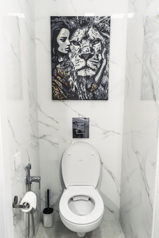 A bathroom with white marble walls and a modern toilet setup. Above the toilet hangs a striking black and white painting featuring a close-up of a lion's face next to a woman's face. The area contains a toilet paper holder with a roll, a toilet brush, and a small handheld bidet.
