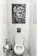 A bathroom with white marble walls and a modern toilet setup. Above the toilet hangs a striking black and white painting featuring a close-up of a lion's face next to a woman's face. The area contains a toilet paper holder with a roll, a toilet brush, and a small handheld bidet.
