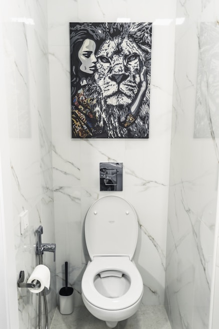 A bathroom with white marble walls and a modern toilet setup. Above the toilet hangs a striking black and white painting featuring a close-up of a lion's face next to a woman's face. The area contains a toilet paper holder with a roll, a toilet brush, and a small handheld bidet.