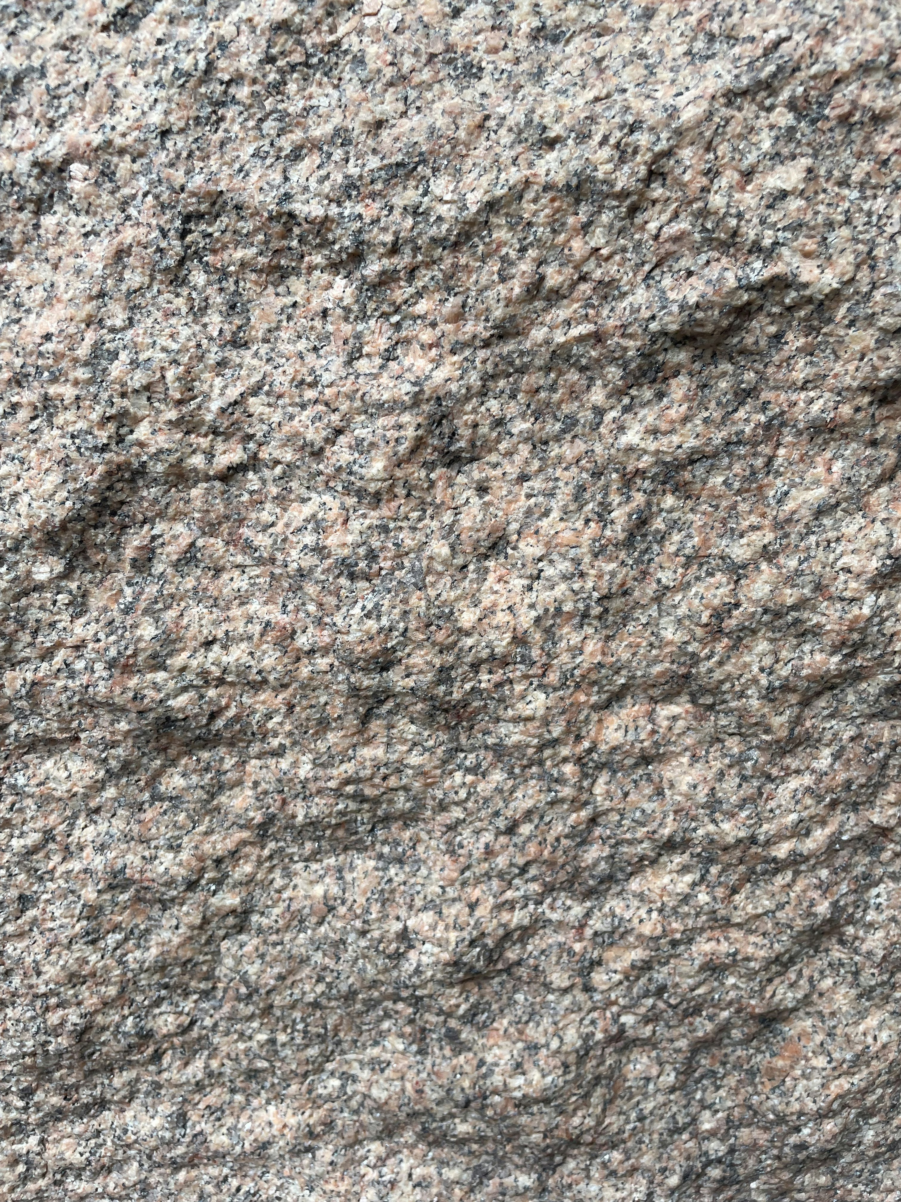 A close-up view of a rough granite surface with a speckled pattern of gray, black, and pinkish hues. The texture is uneven and grainy, indicating natural formations and wear.