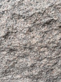 A close-up view of a rough granite surface with a speckled pattern of gray, black, and pinkish hues. The texture is uneven and grainy, indicating natural formations and wear.