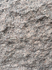 A close-up view of a rough granite surface with a speckled pattern of gray, black, and pinkish hues. The texture is uneven and grainy, indicating natural formations and wear.