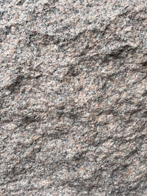 A close-up view of a rough granite surface with a speckled pattern of gray, black, and pinkish hues. The texture is uneven and grainy, indicating natural formations and wear.