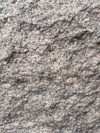 A close-up view of a rough granite surface with a speckled pattern of gray, black, and pinkish hues. The texture is uneven and grainy, indicating natural formations and wear.