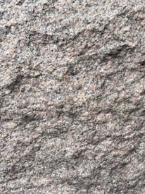 A close-up view of a rough granite surface with a speckled pattern of gray, black, and pinkish hues. The texture is uneven and grainy, indicating natural formations and wear.