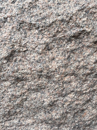 A close-up view of a rough granite surface with a speckled pattern of gray, black, and pinkish hues. The texture is uneven and grainy, indicating natural formations and wear.