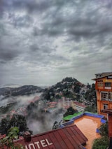 A misty mountainous landscape with buildings spread across the hills. Dense clouds cover the sky, creating a dramatic atmosphere. In the foreground, part of a hotel roof is visible. The scene is lush with greenery and fog, blending the natural environment with urban structures.