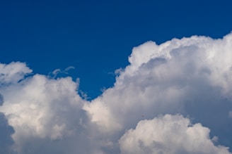 Fluffy white clouds fill most of the image with a deep blue sky as the background. A small parachute is visible high up against the clouds, indicating the presence of a person skydiving.