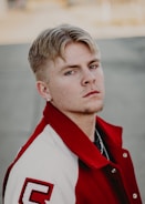 A person with short blond hair and a light mustache is wearing a red and white varsity jacket with a letter 'C' on the sleeve. The background is blurred, focusing attention on the subject's serious expression.
