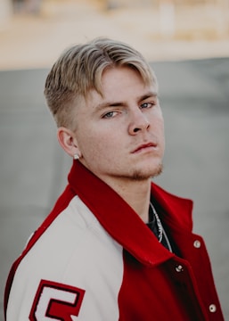 A person with short blond hair and a light mustache is wearing a red and white varsity jacket with a letter 'C' on the sleeve. The background is blurred, focusing attention on the subject's serious expression.