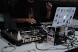 A DJ is performing with a turntable setup. The focus is on the laptop and turntables, with the DJ's hands visible, manipulating the equipment. The laptop is covered with colorful stickers, adding a personal touch. Various cables are connected, contributing to the complexity and industriousness of the scene.