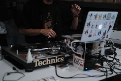 A DJ is performing with a turntable setup. The focus is on the laptop and turntables, with the DJ's hands visible, manipulating the equipment. The laptop is covered with colorful stickers, adding a personal touch. Various cables are connected, contributing to the complexity and industriousness of the scene.