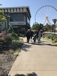 A group of people walking along a paved path toward a large Ferris wheel in an outdoor setting with green trees and landscaped bushes. To the left, a building with a sign reading 'Paula Deen' is visible.