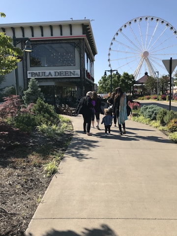 A group of people walking along a paved path toward a large Ferris wheel in an outdoor setting with green trees and landscaped bushes. To the left, a building with a sign reading 'Paula Deen' is visible.