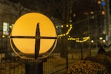 A spherical streetlamp emits a warm, yellow glow, illuminating the surrounding area. The lamp is encased in a decorative metal frame. In the background, a string of small, round lights creates a festive ambiance, softly blurring into a nighttime cityscape with trees and buildings.