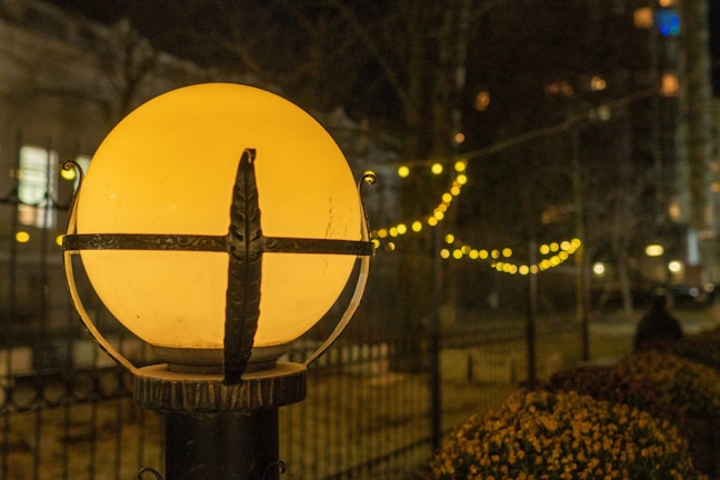 A spherical streetlamp emits a warm, yellow glow, illuminating the surrounding area. The lamp is encased in a decorative metal frame. In the background, a string of small, round lights creates a festive ambiance, softly blurring into a nighttime cityscape with trees and buildings.