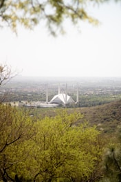 A large, modern mosque is visible in the center of the image, surrounded by lush green hills and trees. The architecture is characterized by a geometric, white structure with tall minarets. The background features a distant view of a city skyline under a hazy sky.