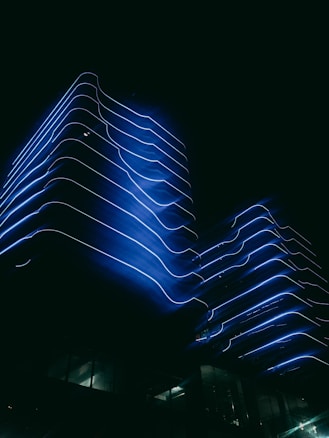A modern building with flowing, wavy blue neon lights outlining its contours against a dark night sky, creating a striking and futuristic appearance. The glow of the lights accentuates the building's architectural design, giving it a surreal and ethereal quality.