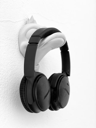 A pair of black over-ear headphones hangs on a uniquely shaped wall mount resembling a face. The wall is white and textured, creating a contrast with the dark color of the headphones.