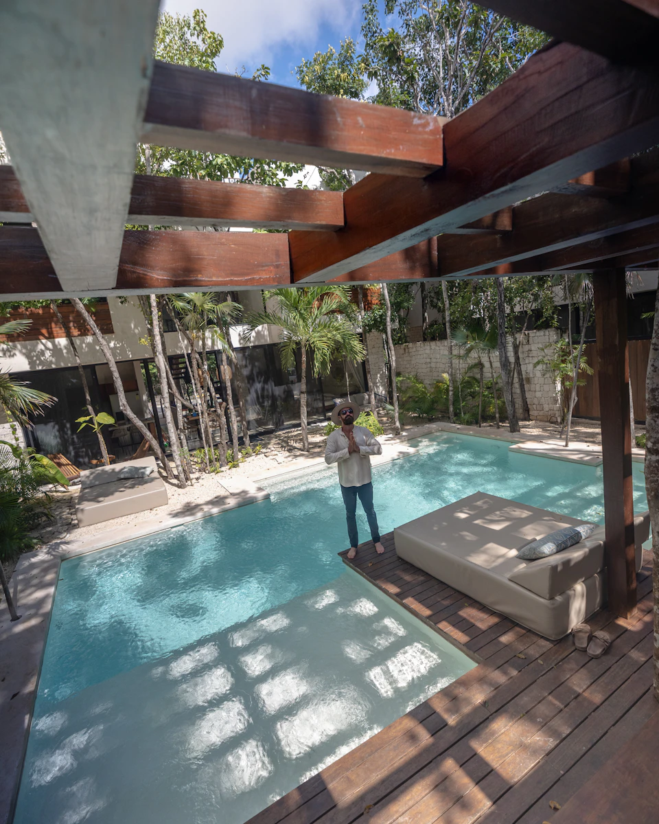 The image depicts a serene outdoor setting with a large pool at its center, surrounded by lush greenery and towering palm trees. The pool is filled with clear blue water that reflects the surrounding environment. A wooden deck extends from the pool area, providing a comfortable space for relaxation or socializing. On one side of the pool, there's a white lounge chair, while on the other side, a person can be seen standing near the edge of the pool.

The scene is captured from an elevated perspective, offering a bird's eye view of the entire area. The image also contains some text that reads "sa_20".