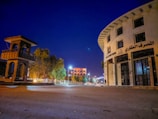 A wide street scene at night features a distinctive round building with multiple stories labeled in Arabic and English as 'Real Estate Bank.' On the left, there's a tall, open-structured stone tower. Streetlights illuminate the area, casting a soft glow. Trees line the sidewalk, and a few modern buildings are visible in the background under the clear deep blue sky.
