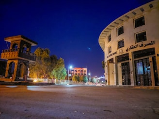 A wide street scene at night features a distinctive round building with multiple stories labeled in Arabic and English as 'Real Estate Bank.' On the left, there's a tall, open-structured stone tower. Streetlights illuminate the area, casting a soft glow. Trees line the sidewalk, and a few modern buildings are visible in the background under the clear deep blue sky.