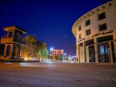 A wide street scene at night features a distinctive round building with multiple stories labeled in Arabic and English as 'Real Estate Bank.' On the left, there's a tall, open-structured stone tower. Streetlights illuminate the area, casting a soft glow. Trees line the sidewalk, and a few modern buildings are visible in the background under the clear deep blue sky.
