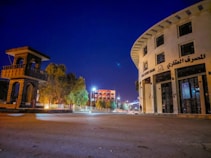 A wide street scene at night features a distinctive round building with multiple stories labeled in Arabic and English as 'Real Estate Bank.' On the left, there's a tall, open-structured stone tower. Streetlights illuminate the area, casting a soft glow. Trees line the sidewalk, and a few modern buildings are visible in the background under the clear deep blue sky.
