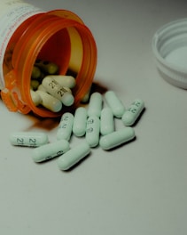 Several white capsules with numbers are scattered across a surface, with an orange prescription bottle lying on its side. The pills spill out of the container, and a white bottle cap is visible nearby.