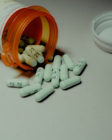 Several white capsules with numbers are scattered across a surface, with an orange prescription bottle lying on its side. The pills spill out of the container, and a white bottle cap is visible nearby.