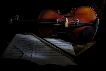 A violin rests on a dark surface with sheet music scattered beneath it. The lighting is low, casting shadows that create a dramatic and intimate atmosphere. The violin, a classic wooden instrument, appears well-used, with a rich reddish-brown hue. The bow lies across the sheets of music, accentuating the theme of musicality.