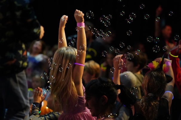 Children are gathered in a dark environment, with one child in the foreground raising arms surrounded by bubbles. Neon bracelets and colorful outfits suggest a festive atmosphere. Multiple children, some of whom seem to be playing, are involved in what looks like a party or event.