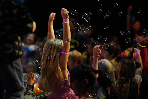 Children are gathered in a dark environment, with one child in the foreground raising arms surrounded by bubbles. Neon bracelets and colorful outfits suggest a festive atmosphere. Multiple children, some of whom seem to be playing, are involved in what looks like a party or event.
