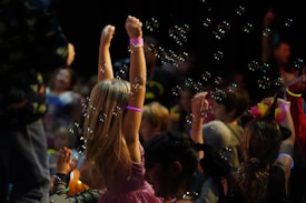 Children are gathered in a dark environment, with one child in the foreground raising arms surrounded by bubbles. Neon bracelets and colorful outfits suggest a festive atmosphere. Multiple children, some of whom seem to be playing, are involved in what looks like a party or event.