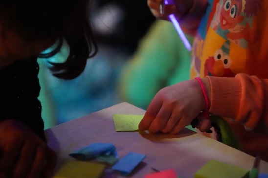 A child, partially visible, is engaging in a creative activity, holding a pen and drawing on a piece of yellow paper. The child is wearing a colorful shirt with cartoon characters on it. Various colored papers are scattered on the table in front of them, suggesting an art or crafting environment. The lighting is dim, with a focus on the vibrant colors of the paper and shirt.