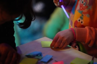 A child, partially visible, is engaging in a creative activity, holding a pen and drawing on a piece of yellow paper. The child is wearing a colorful shirt with cartoon characters on it. Various colored papers are scattered on the table in front of them, suggesting an art or crafting environment. The lighting is dim, with a focus on the vibrant colors of the paper and shirt.