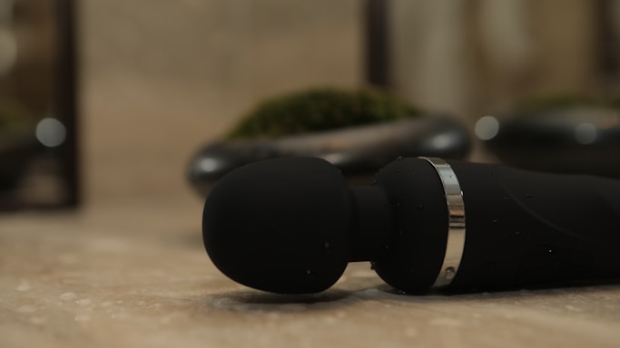 A close-up view of a black handheld massager lying on a smooth, beige surface. The massager has a round head and a metallic ring near its base. In the blurred background, there are some decorative elements, possibly small plants in pots.
