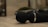A close-up view of a black handheld massager lying on a smooth, beige surface. The massager has a round head and a metallic ring near its base. In the blurred background, there are some decorative elements, possibly small plants in pots.