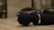 A close-up view of a black handheld massager lying on a smooth, beige surface. The massager has a round head and a metallic ring near its base. In the blurred background, there are some decorative elements, possibly small plants in pots.