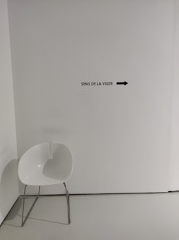 A minimalist setting with a modern white chair featuring metal legs placed in a corner. A wall sign reads 'Sens de la visite' with an arrow pointing right.