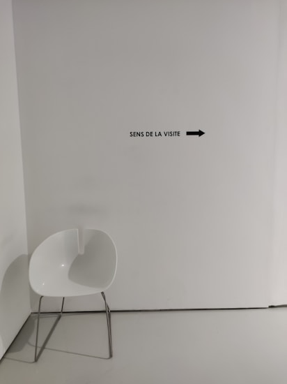 A minimalist setting with a modern white chair featuring metal legs placed in a corner. A wall sign reads 'Sens de la visite' with an arrow pointing right.